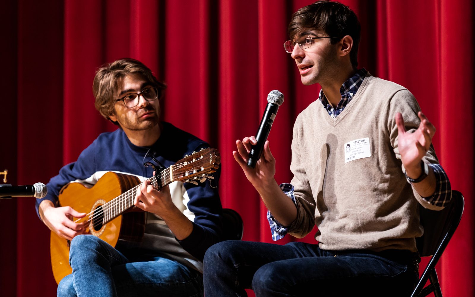 Daniel Lazour with a guitar and Patrick Lazour with a microphone onstage in front of red curtain.