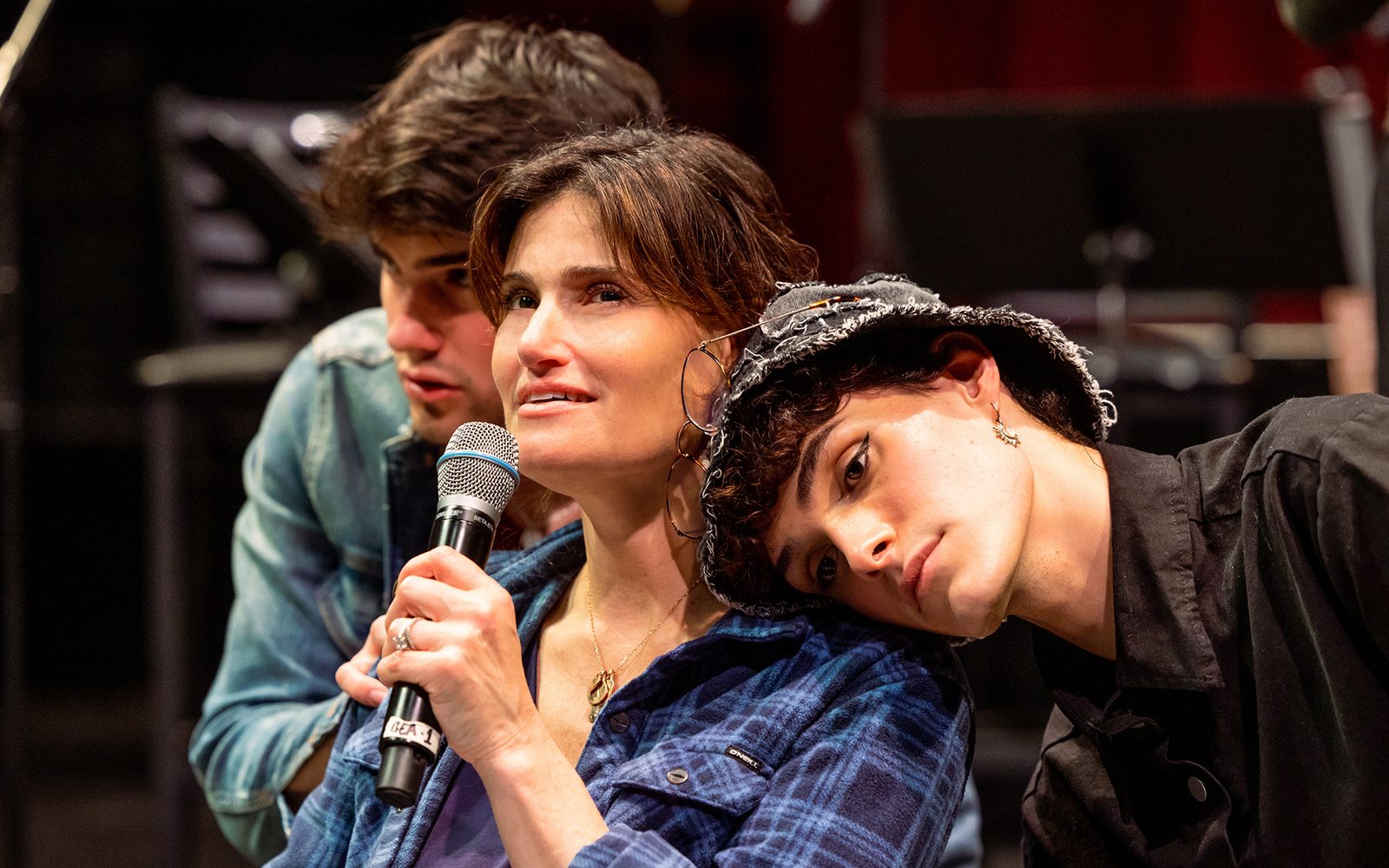 Idina Menzel speaking into a microphone while Michael Williams and Luke Ferrari lean on her.