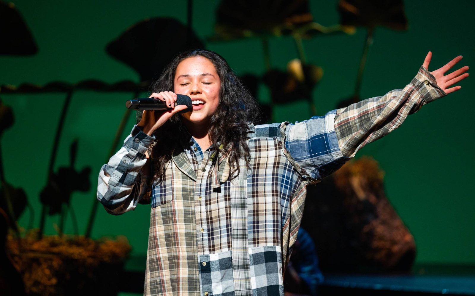YDE singing into a mic with her arm flung open in WILD: A Musical Becoming.