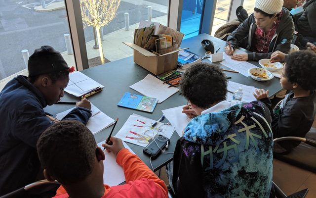 A group of youths draw at a table scattered with papers.