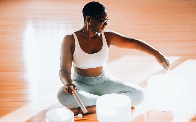 A woman sitting on a sun-strewn floor playing singing bowls.