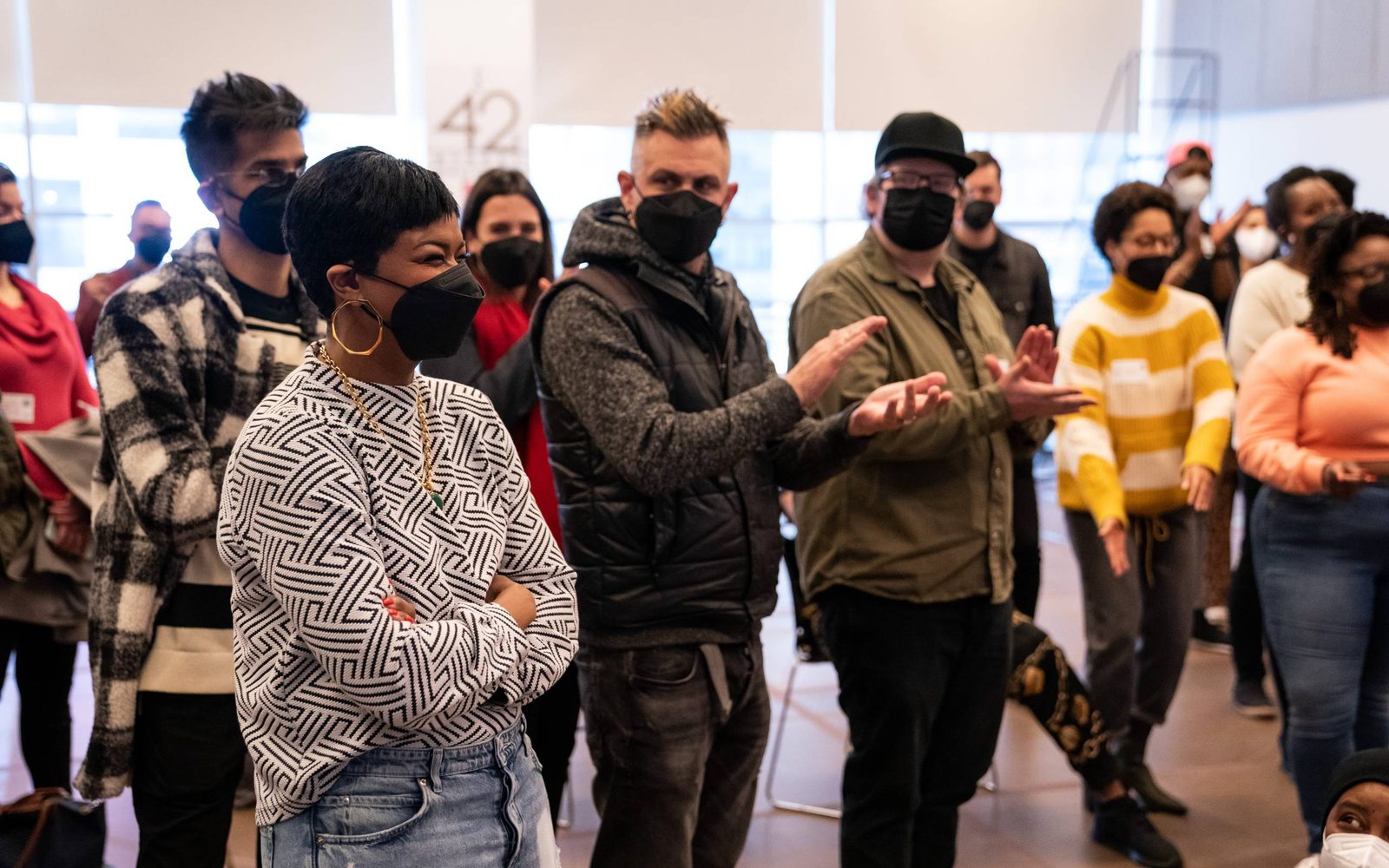 Hair, Make-Up, and Wig Designer Mia Neal with members of the company behind her clapping.