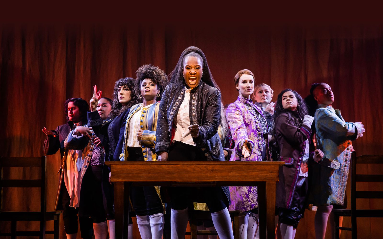 Sushma Saha, Sara Porkalob, Mehry Eslaminia, Gisela Adisa, Elizabeth A. Davis, Becca Ayers, Brooke Simpson, and Oneika Phillips arranged in V behind Crystal Lucas-Perry and a desk.