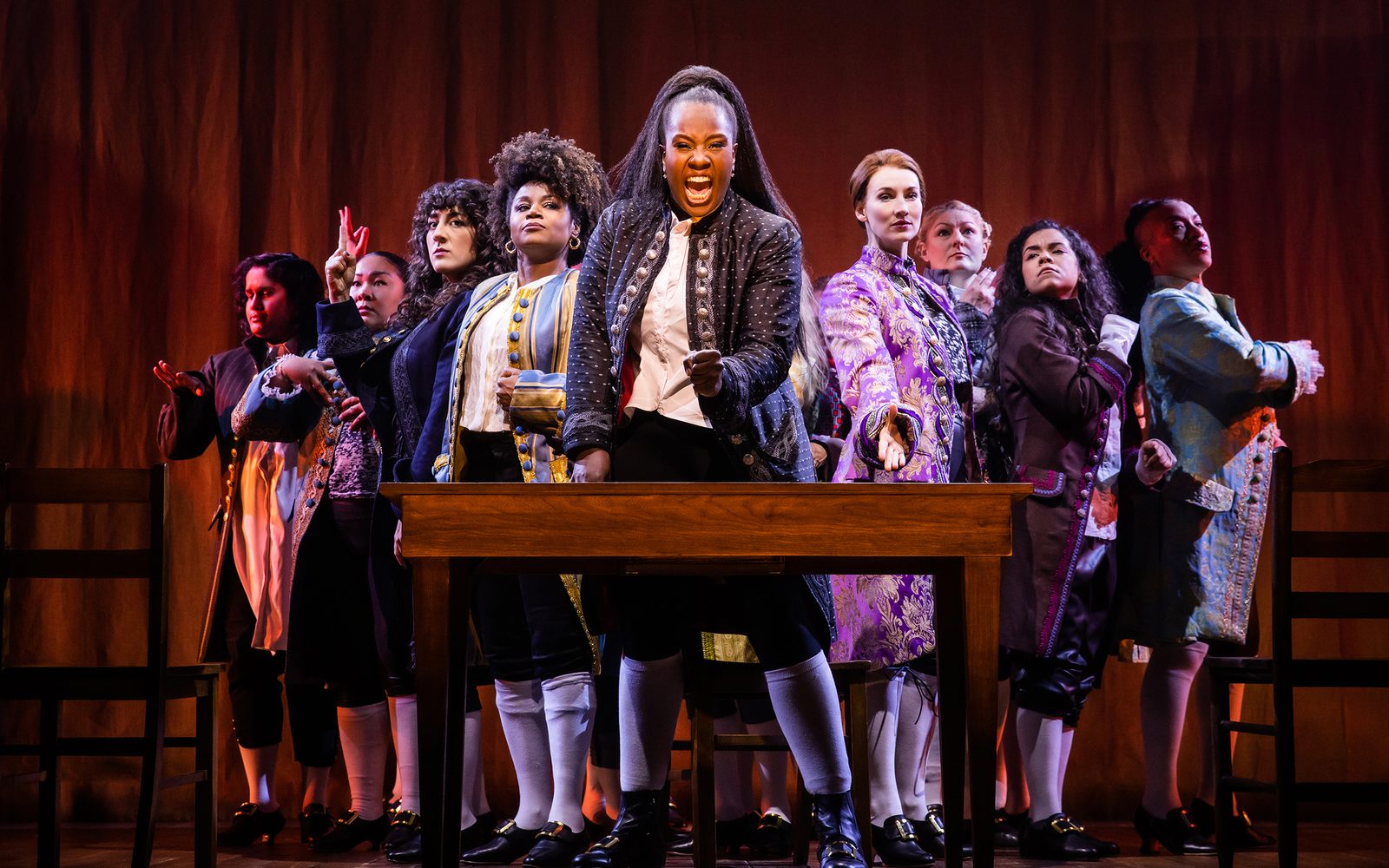 Sushma Saha, Sara Porkalob, Mehry Eslaminia, Gisela Adisa, Elizabeth A. Davis, Becca Ayers, Brooke Simpson, and Oneika Phillips arranged in V behind Crystal Lucas-Perry and a desk.