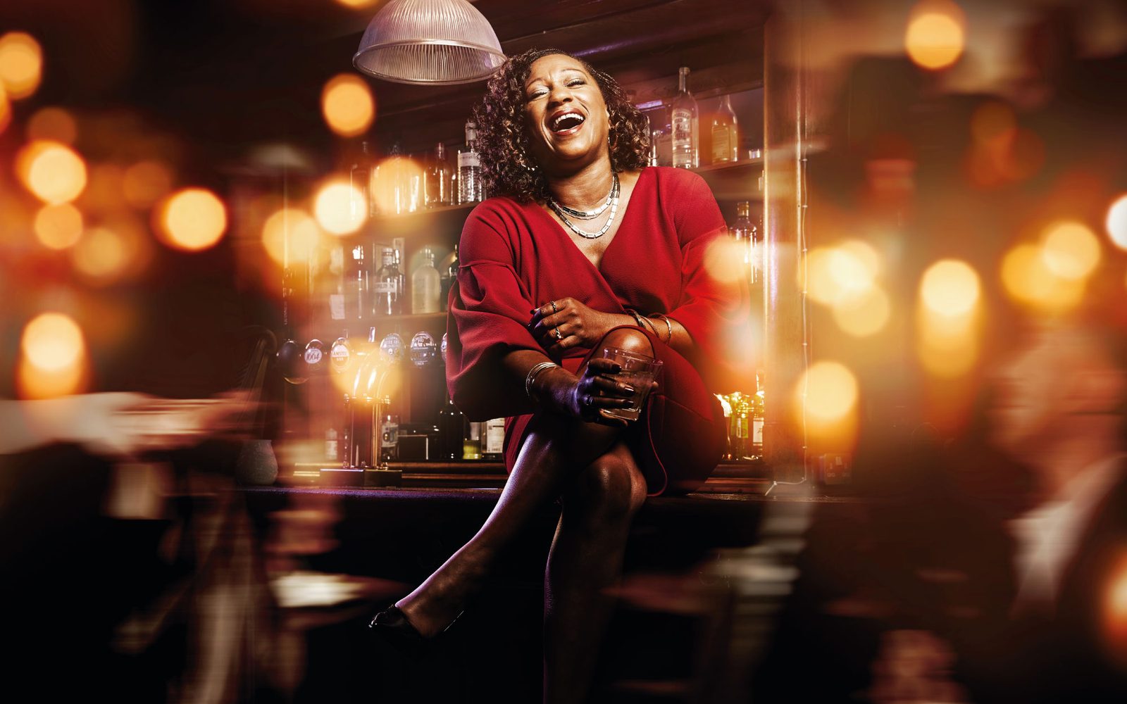 Woman in red dress sits atop a bar laughing with a scotch glass in hand.