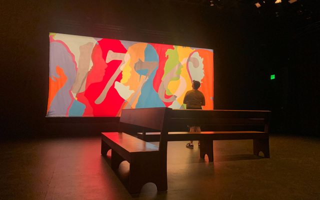 A person n front of a bench stares at a large, colorful mural in a dark room.