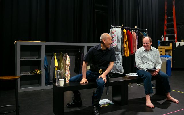 Francis Jue and Carl Palmer smile while talking to another while sitting on a bench in rehearsal.