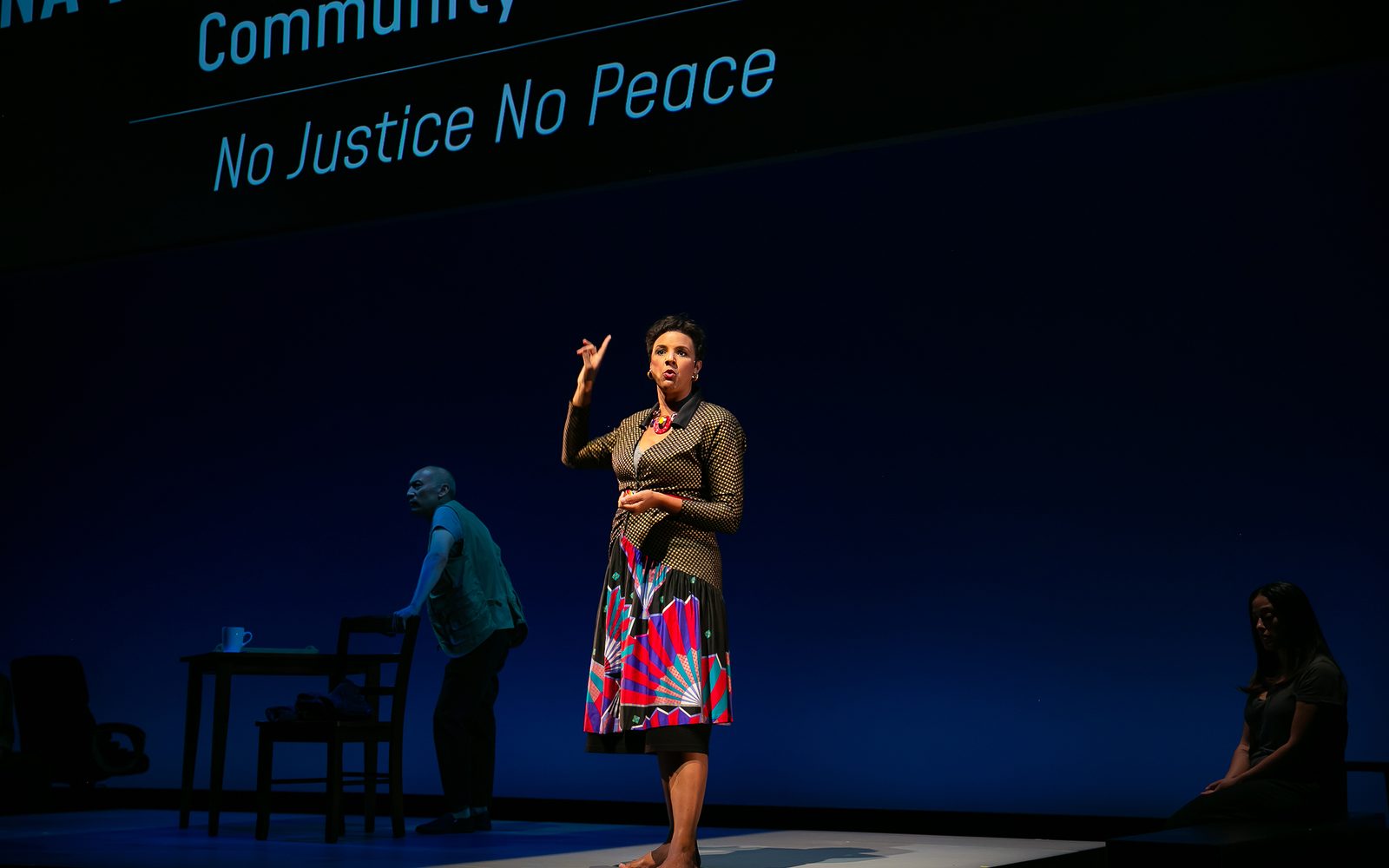 Tiffany Rachelle Stewart in a checkered blazer and printed skirt pointing up, in front of a wall that says, in part, “No Justice No Peace”.