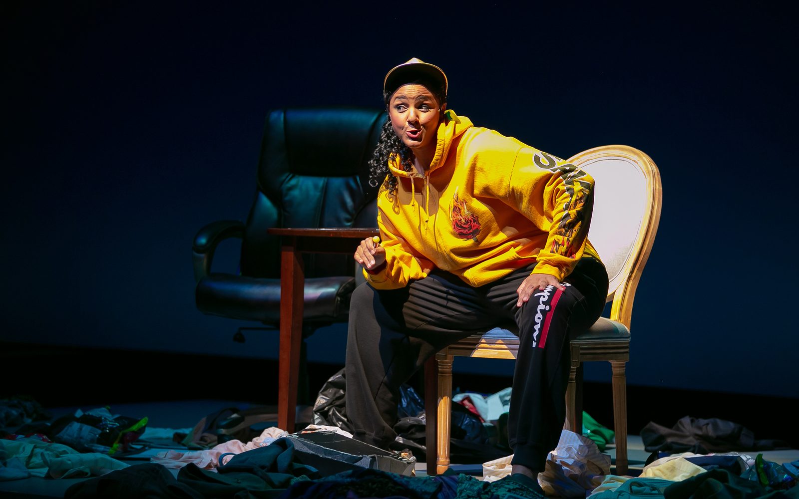 Tiffany Rachelle Stewart in a yellow sweatshirt, black sweatpants, and baseball cap sitting amidst a piles of clothes.