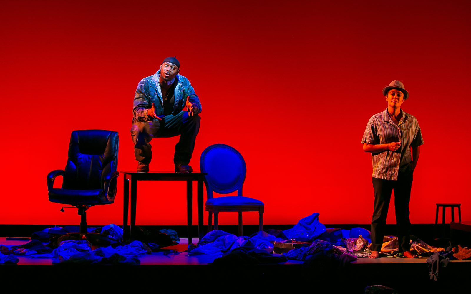 Wesley T. Jones in a patched denmic jacket, jeans, and a durag squats on a table between chairs, while Elena Hurst in a striped button-down and fedora stands to the right.