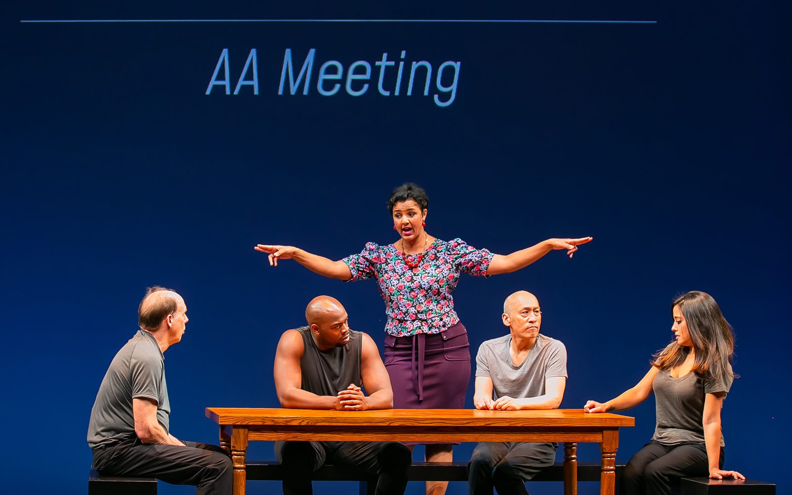 Carl Palmer, Wesley T. Jones, Francis Jue, and Elena Hurst sit at a table while Tiffany Rachelle Hurst gestures broadly in front a wall that says, “The Words of Maria, Juror #7, Federal Trial: ‘AA Meeting’”.