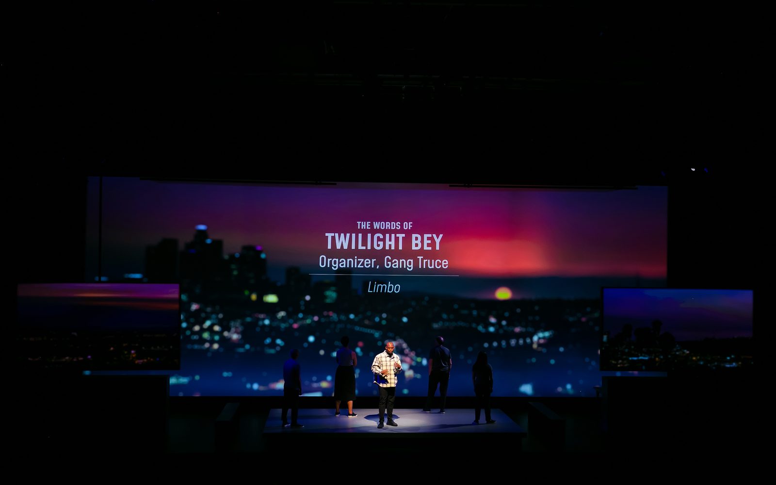 Wesley T. Jones spotlit on a darkened stage, in front of dusky cityscape that says, “The Words of Twilight Bey, Organizer, Gang Truce: ‘Limbo’”.