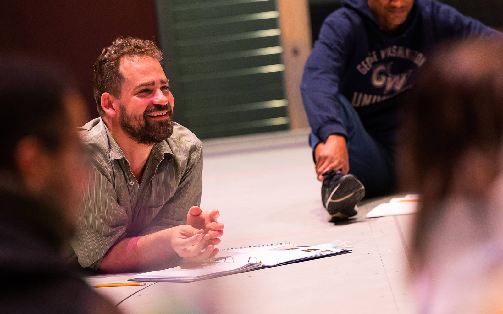 Max Webster lying on the ground in front of an open script between cast members sitting on the ground.