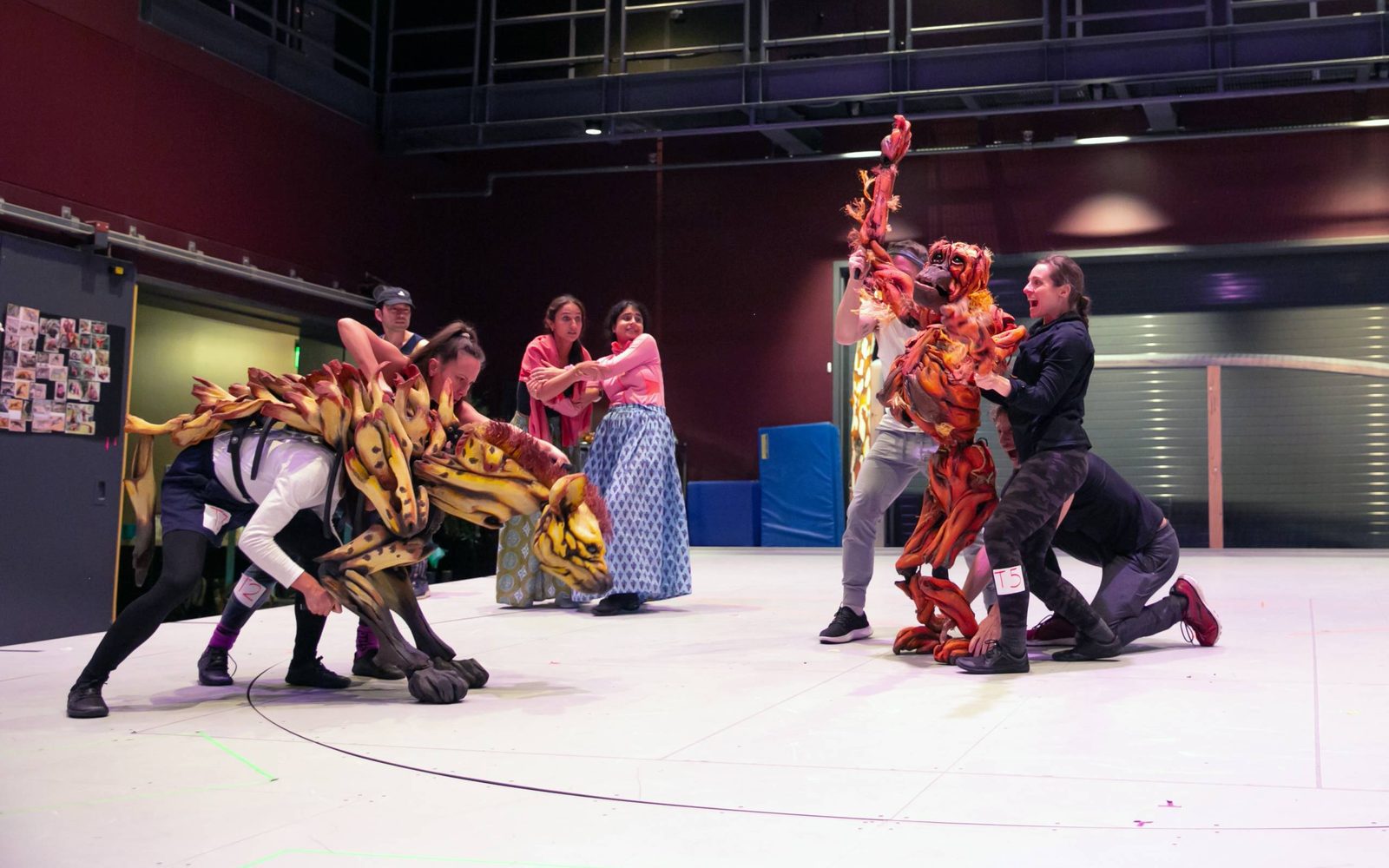 Celia Mei Rubin manipulates the front of a hyena puppet with Nikki Calonge handling its rear in front of Andrew Wilson, Jonathan David Martin, and Betsy Rosen holding up an orangutan puppet, while Sonya Venugopal and Mahira Kakka look in fear between them.