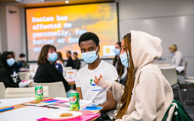 A student with braids falling out of a hoodie speaks while another looks on, in a room of people sitting at tables.
