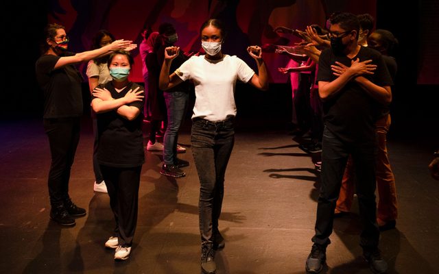 Two groups of a people stand in lines with their arms in different gestures, while another stands in the middle with her firsts raised.