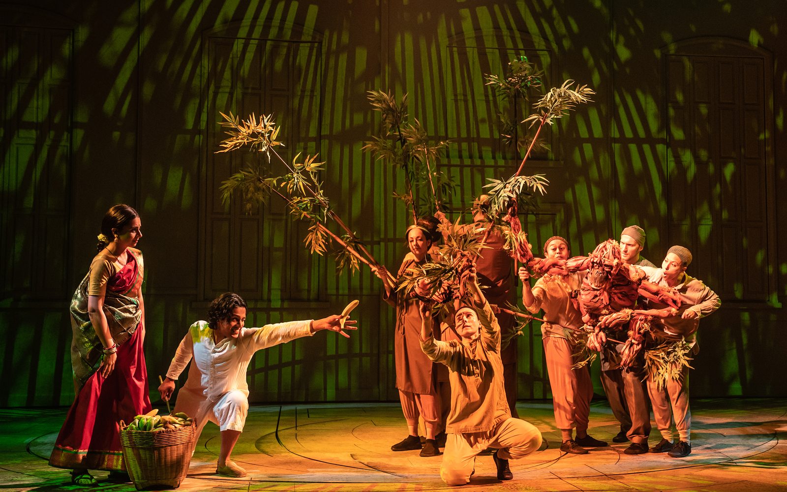Mahira Kakkar watches Adi Dixit reach out a banana towards Salma Shaw, Scarlet Wilderink, Nikki Calonge, Rowan Magee, and Celia Mei Rubin manipulating an orangutan puppet through bamboo stalks.