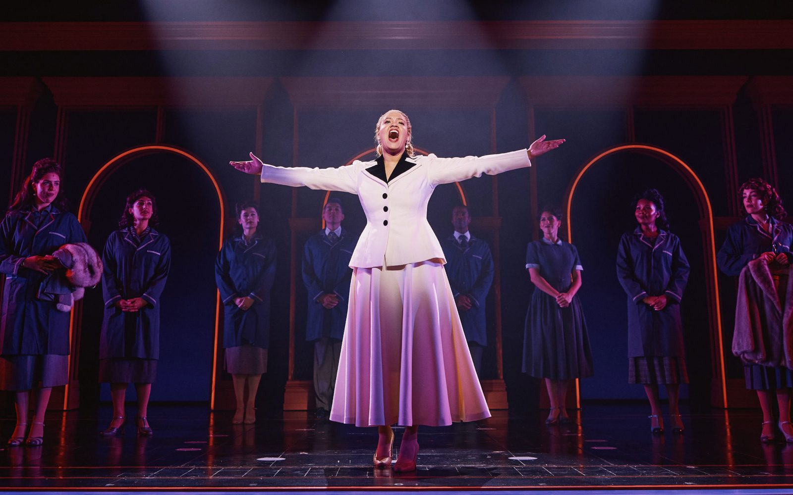 Shereen Pimentel as Eva standing in the spotlight with her arms outstretched while cast members look on.