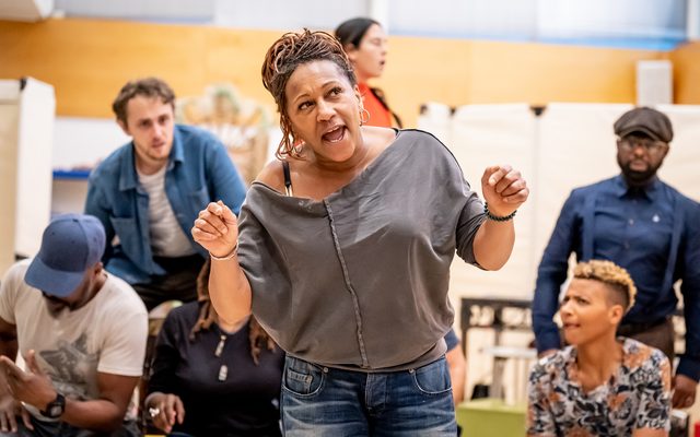 Clare Perkins gesticulating and speaking with the rest of the company behind her watching.