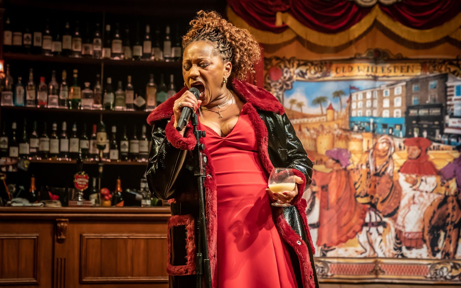 Clare Perkins in a red dress and fur-lined leather jacket, shouts into a microphone with one hand and holds a glass of brandy in the other.