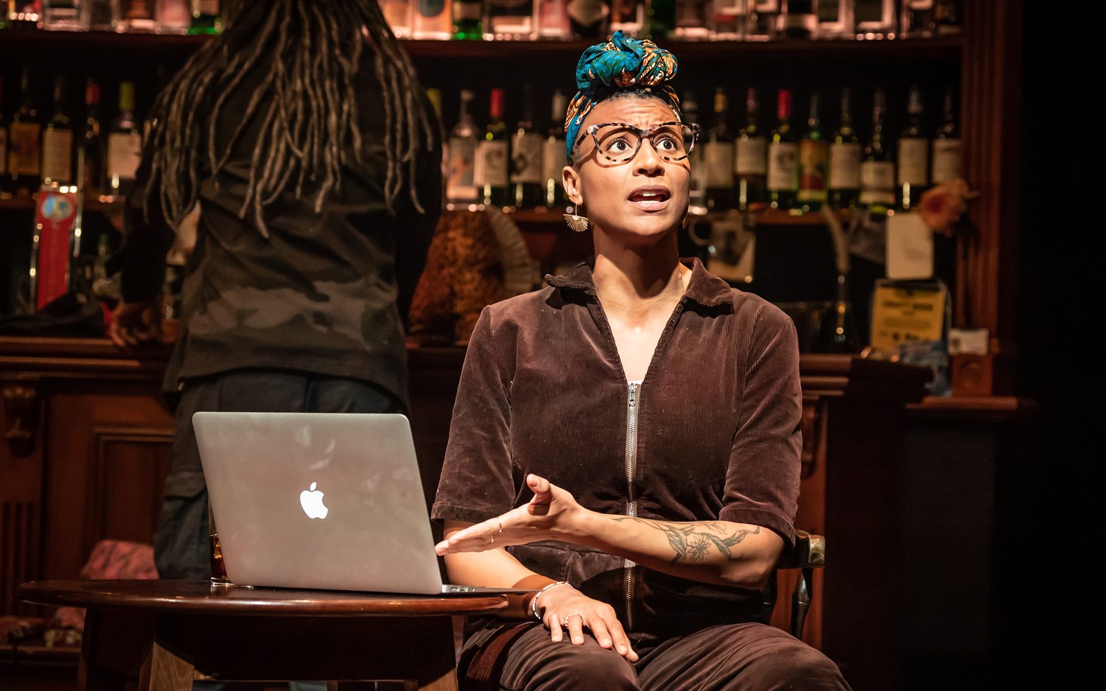Jessica Murrain in a colorful head wrap, tortoiseshell glasses, and burgundy corduroy jumpsuit sitting at a small table motioning to a laptop on it.