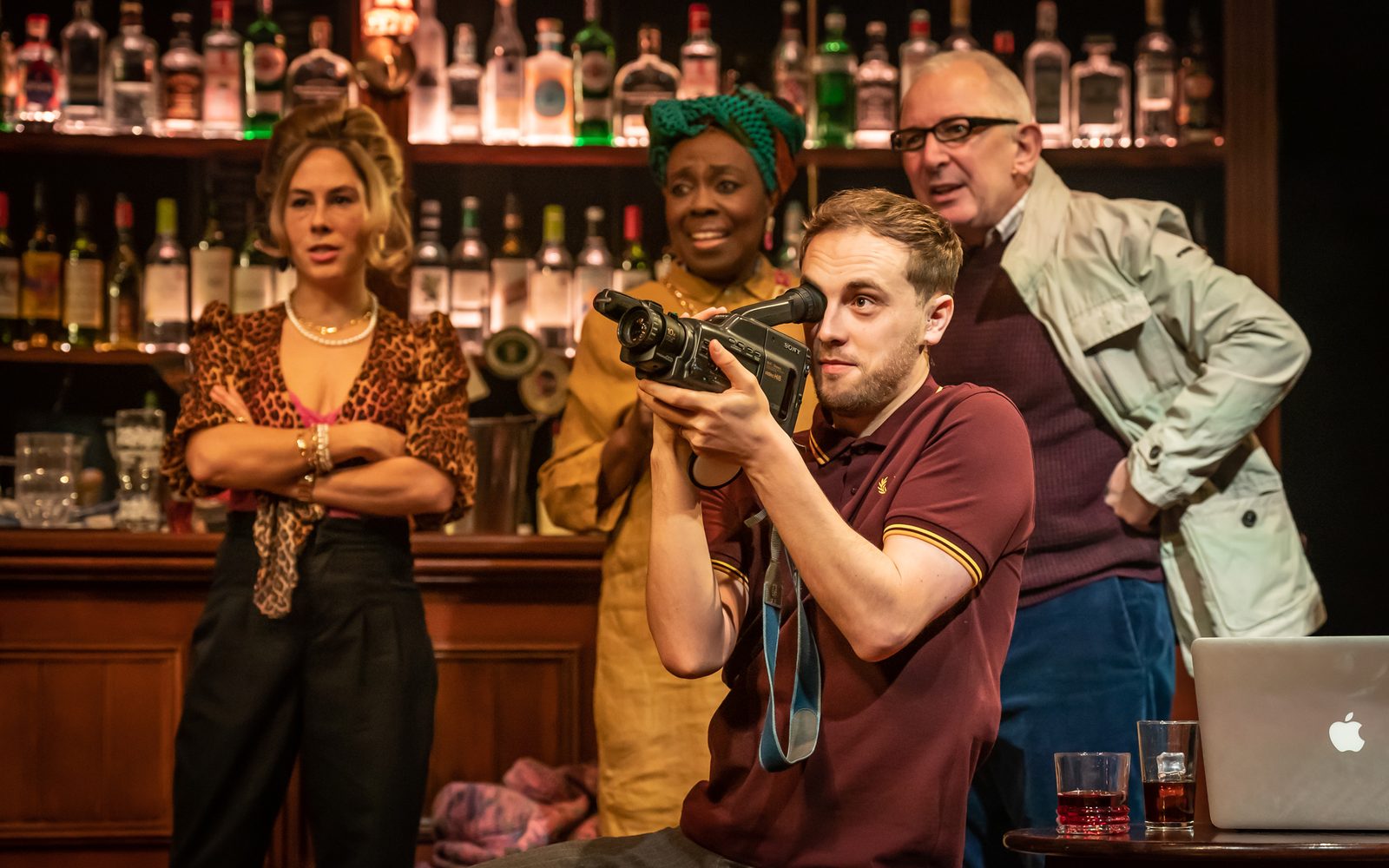 Scott Miller holds a hand-held camcorder to his eye, pointing to the left, while Claudie Grant, Ellen Thomas, and Andrew Frame look on from the bar behind him.
