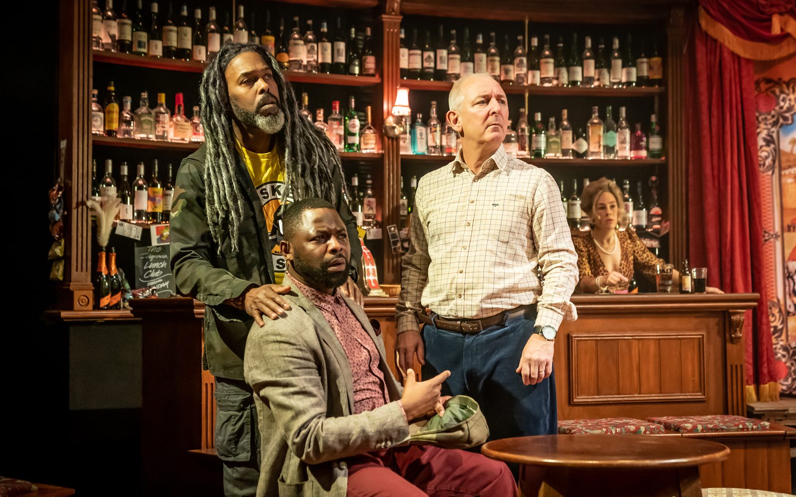 Marcus Adolphy and Andrew Frame stand behind George Eggay, who sits at a table, looking worriedly at something to the right. Claudia Grant stands behind the bar to the right, also staring.