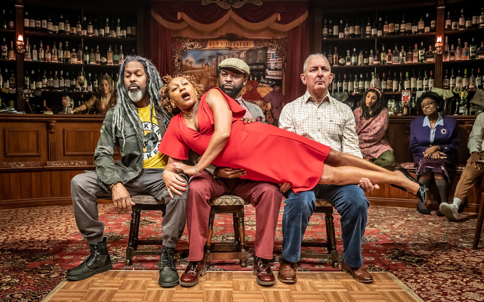 Marcus Adolphy, George Eggay, and Andrew Frame sit on stools in a line while Clare Perkins lies on top of their laps.