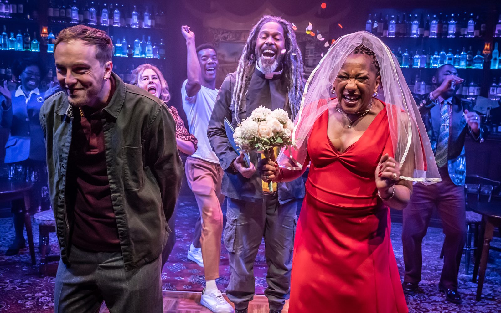 Scott Miller on the left has a wide grin, while Clare Perkins in a white veil and holding a bouquet of white flowers laughs. Behind them, Marcus Adolphy in a clerical collar and the company shout in celebration.