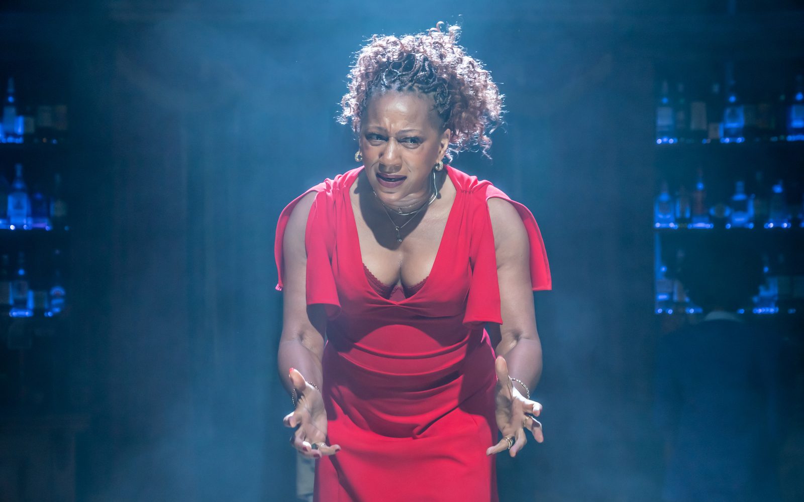 Clare Perkins in a red dress and bather in eery light and fog, leans forward with her hands outstretched.