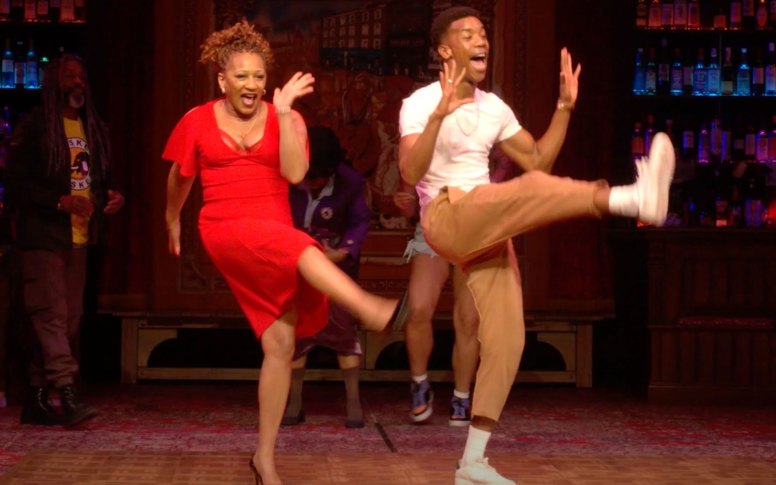 Clare Perkins in a red dress and Troy Glasgow in a white T and khaki joggers do a high kick while dancing