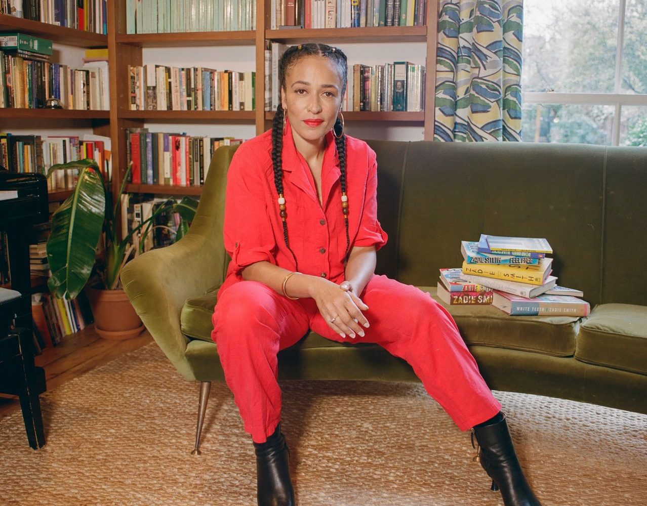Zadie Smith in a red jumpsuit seated on a muted green couch in front of a bookshelf in her living room.