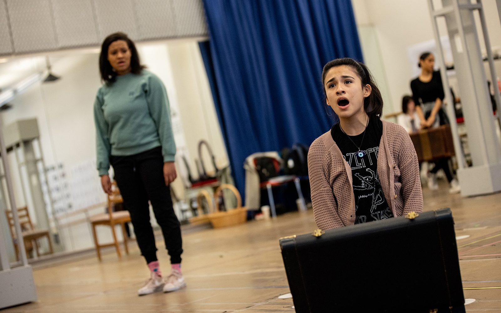 Naomi Serrano sings kneeled in front of an open old-fashioned leather suitcase while Shereen Pimentel stands at a distance watching her.