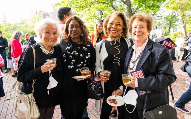 Four women dressed to impress stand together smiling on a sunny outdoor patio.