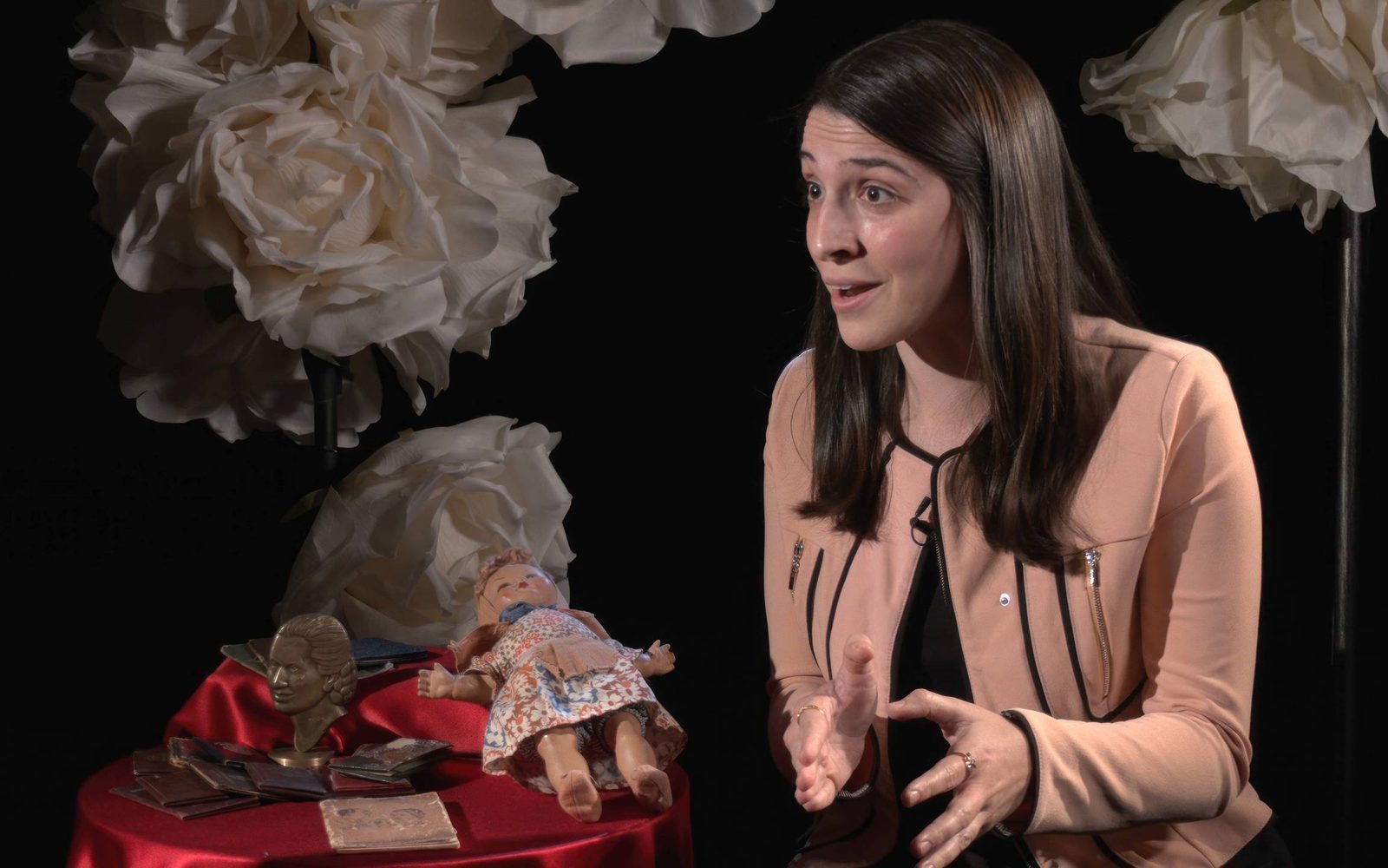 Director Sammi Cannold beside a table with items pertaining to Eva Perón: a doll, a booklet, and a small bust.