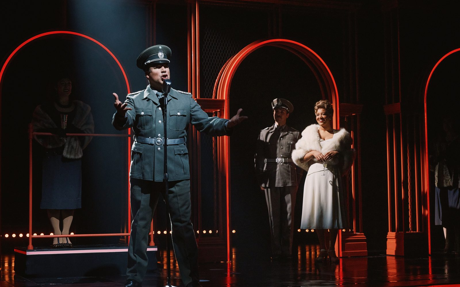 Caesar Samayoa as Perón speaks into a microphone, gesturing dramatically while Caleb Marshall-Villarreal, and Shereen Pimentel (Eva) look on.