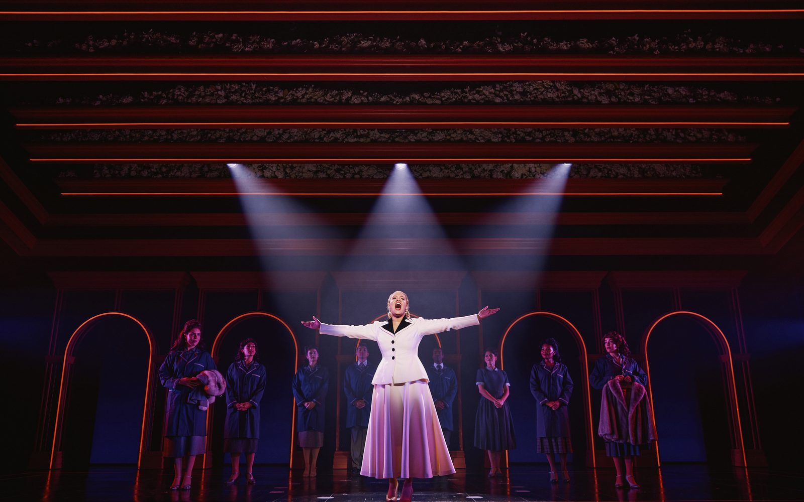 Shereen Pimentel as Eva standing in the spotlight with her arms outstretched while cast members look on.