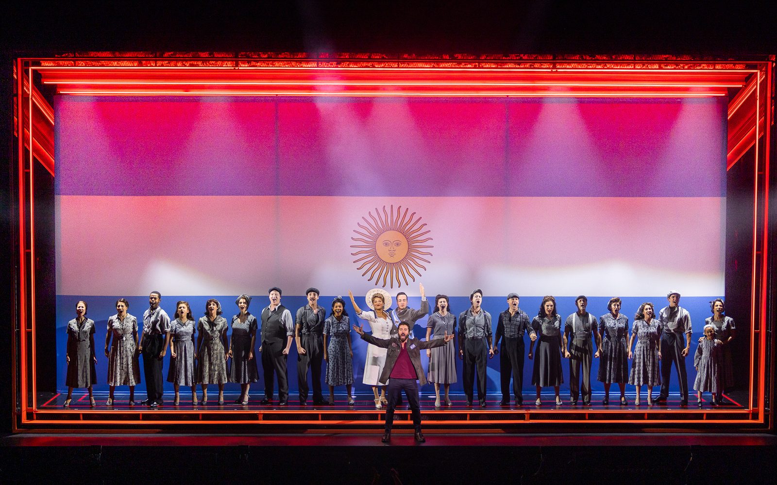 The Company of Evita stands in front of a large flag of Argentina.