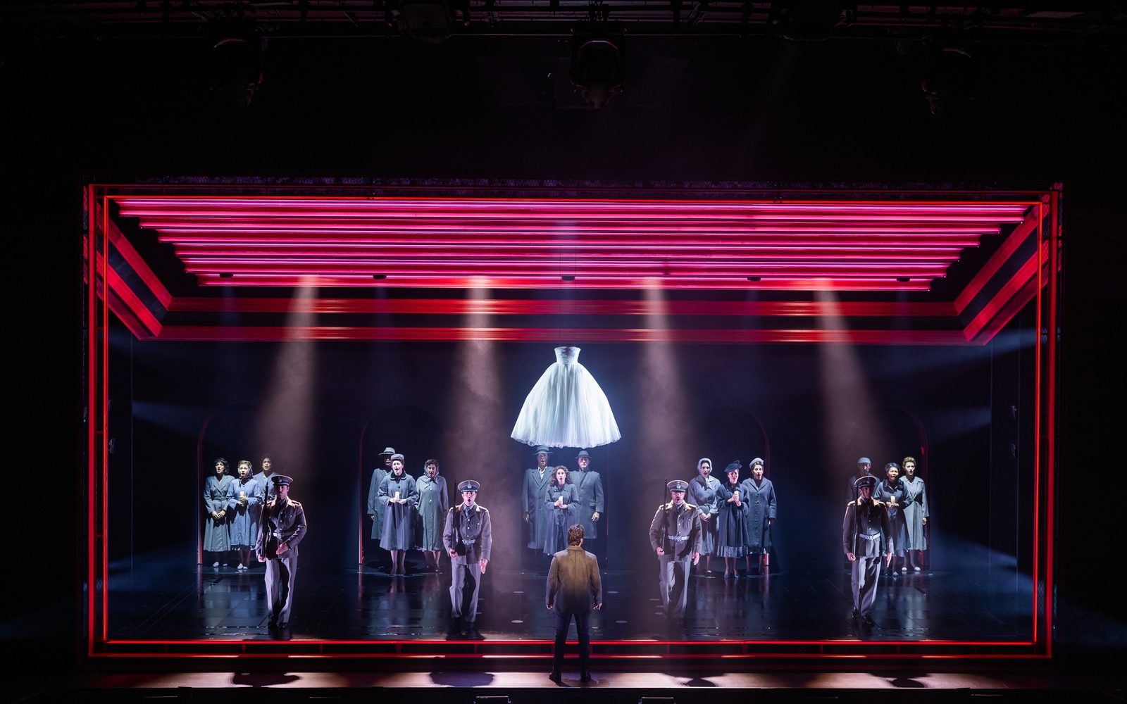 The company of Evita sings on stage, standing in small groups. They are framed by red lights and a white dress hangs overhead.