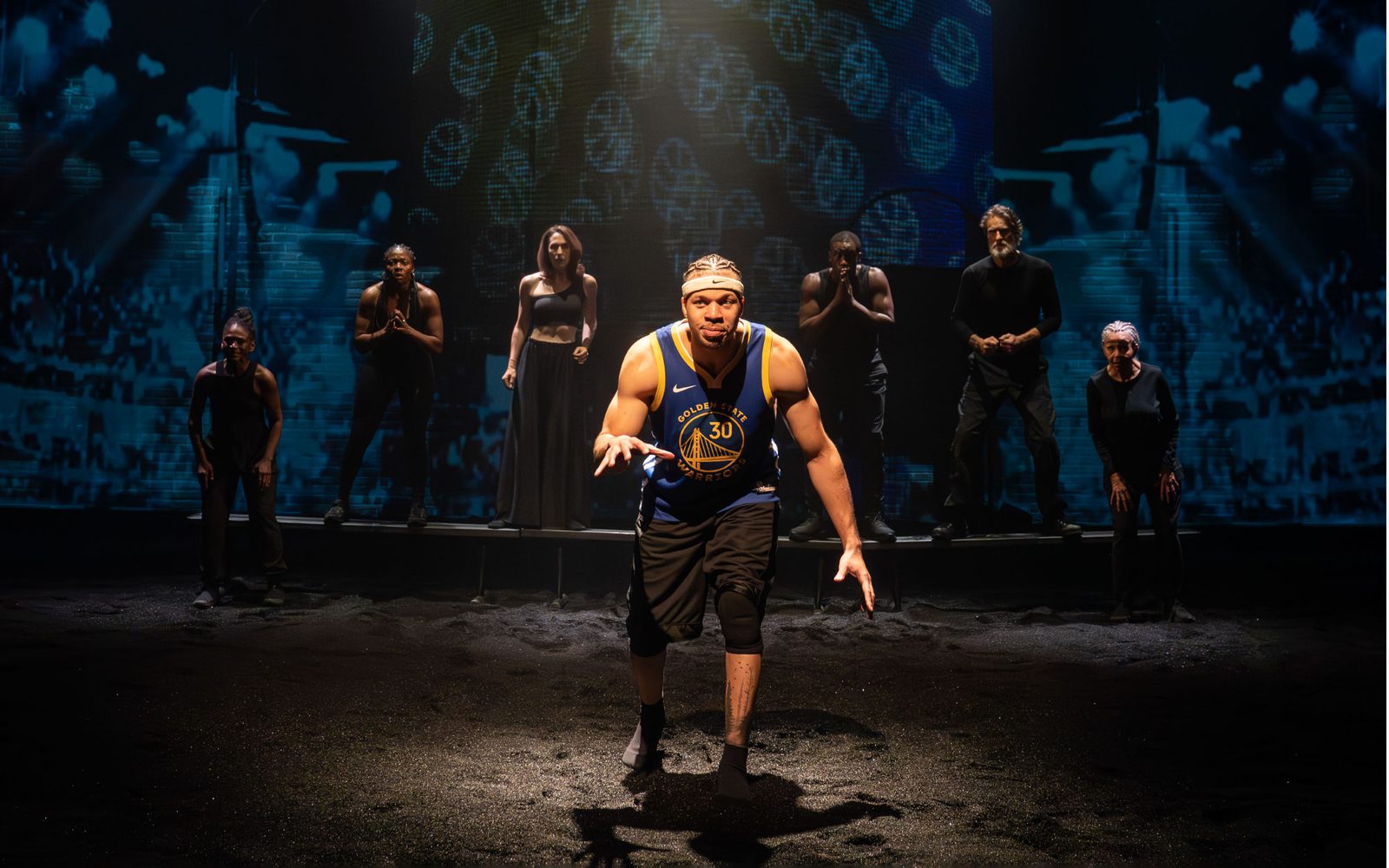 Mister Fitzgerald in a Lakers jersey is spotlit in front of the rest of the cast, lined behind him and in black.