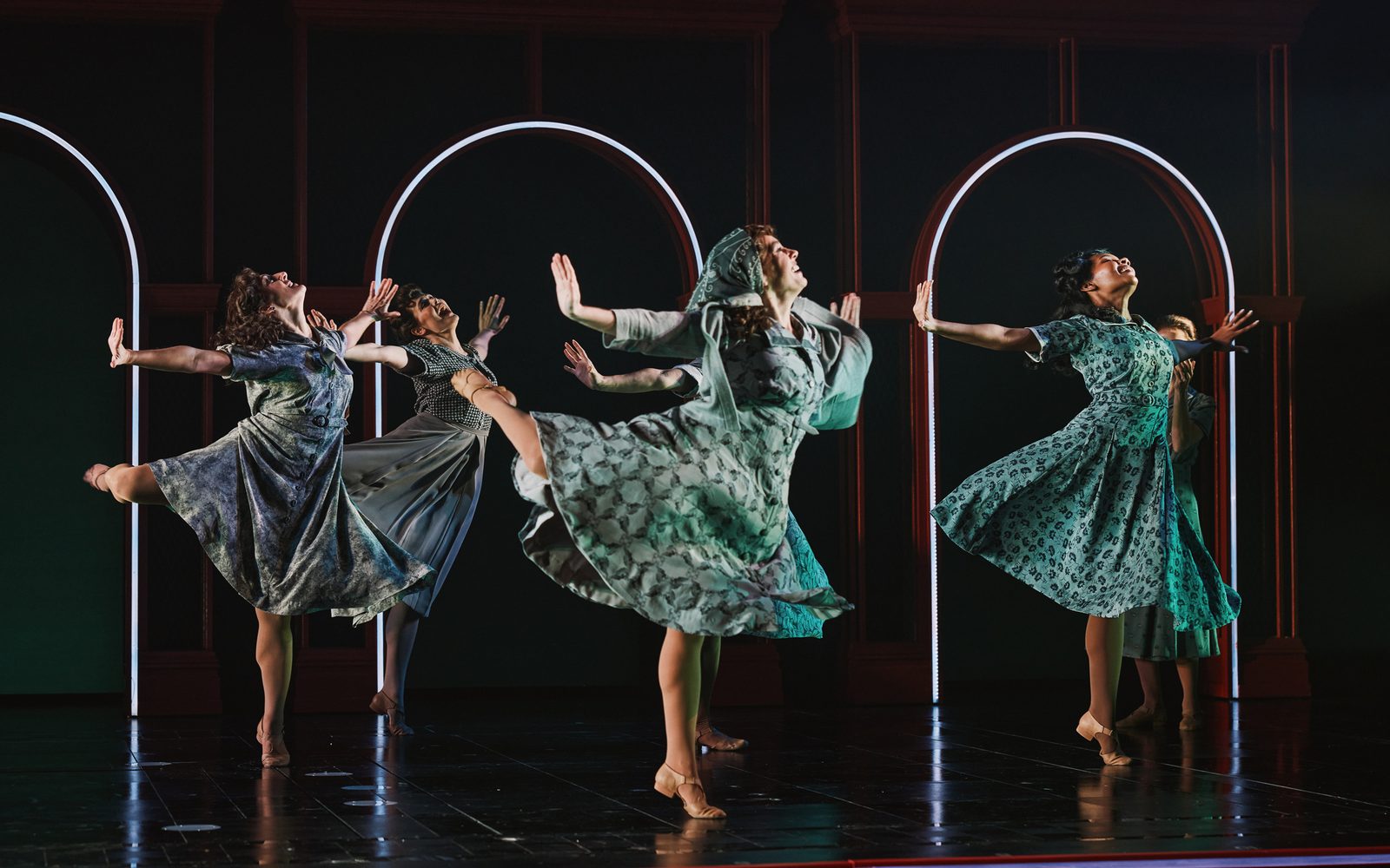 Ensemble members dance on stage with arms outstretched and one leg lifted in the air.