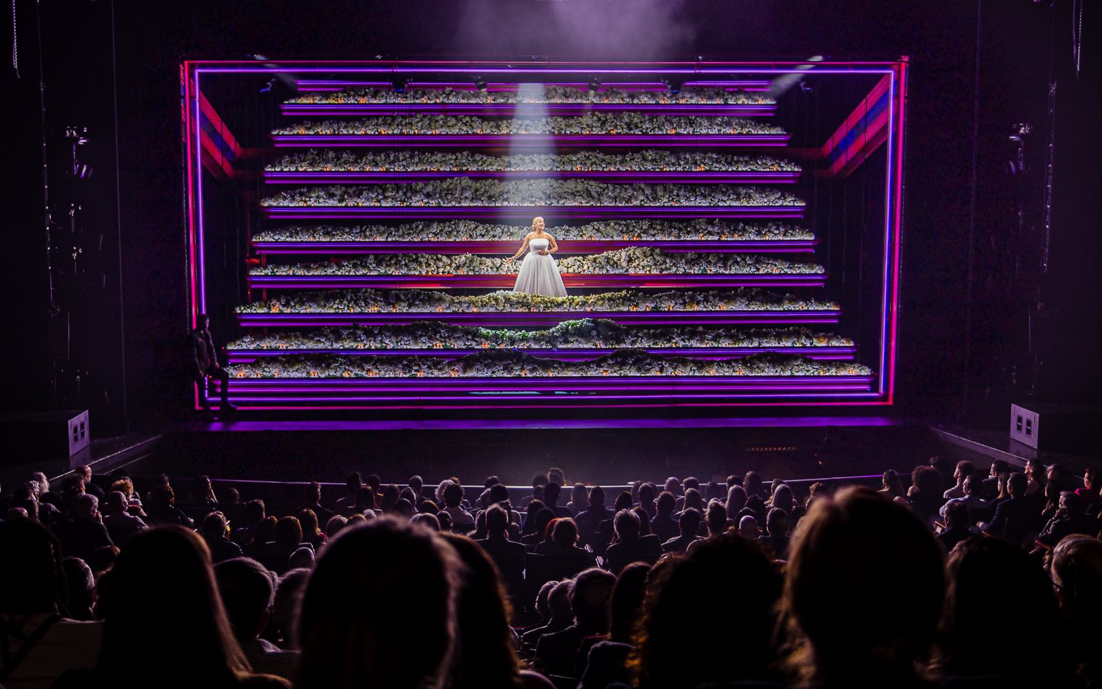 Shereen Pimentel as Eva standing on stage on steps covered in white flowers while the audience looks on.