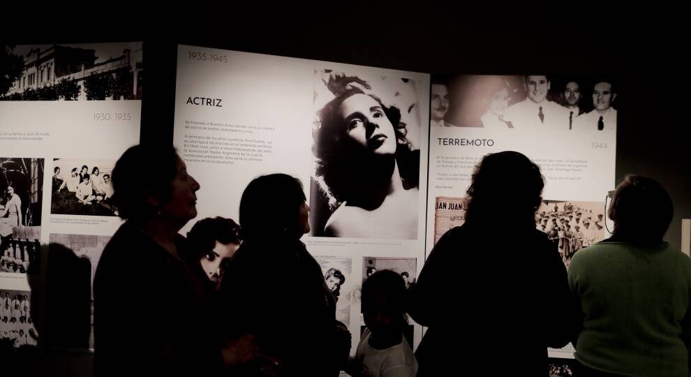 Silhouettes of people in front of a museum display featuring black and white images of Eva Peron, news clippings, and historic images.