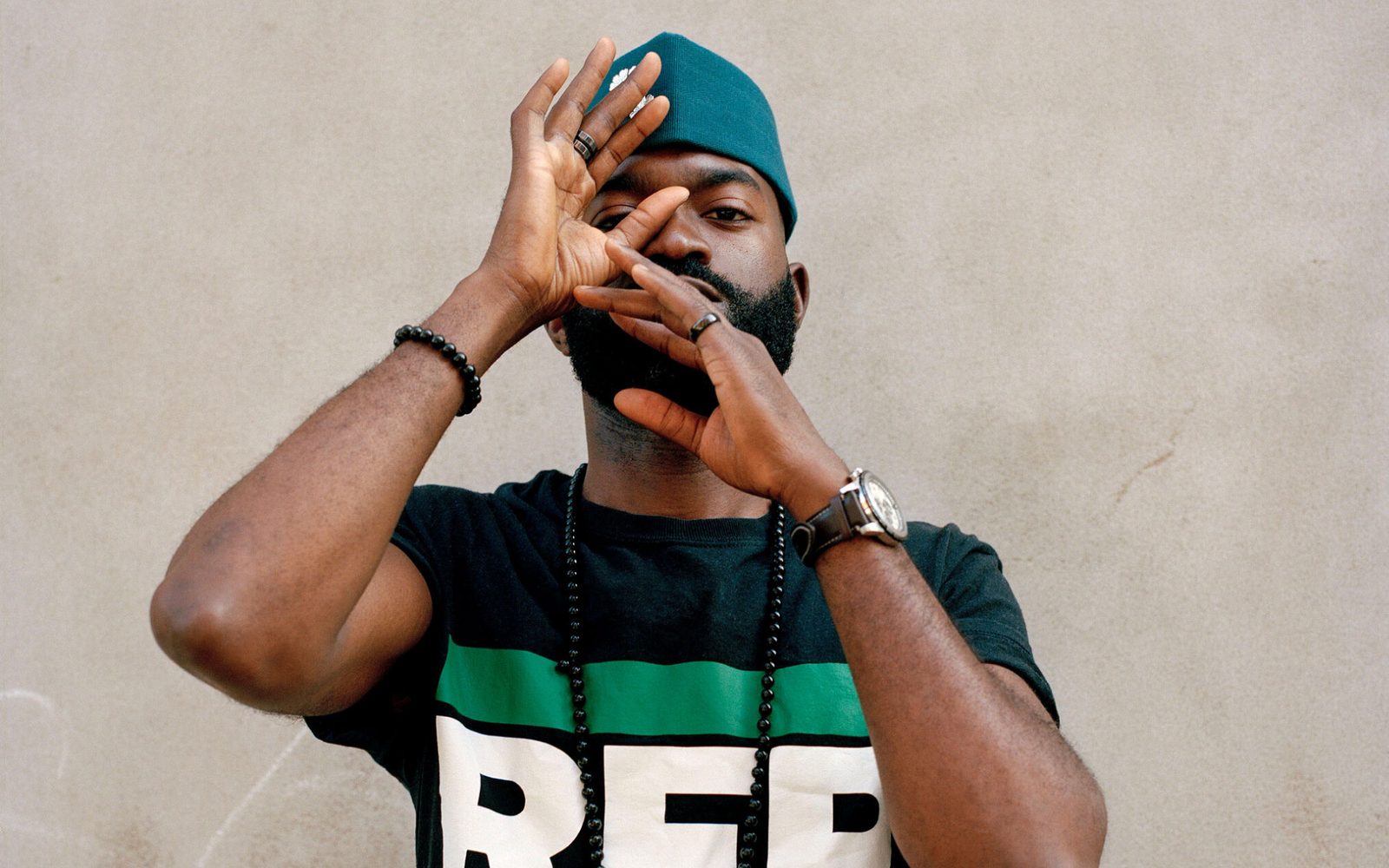 Inua Ellams in a kufi hat and graphic T-shirt, raises his hands up to his face.