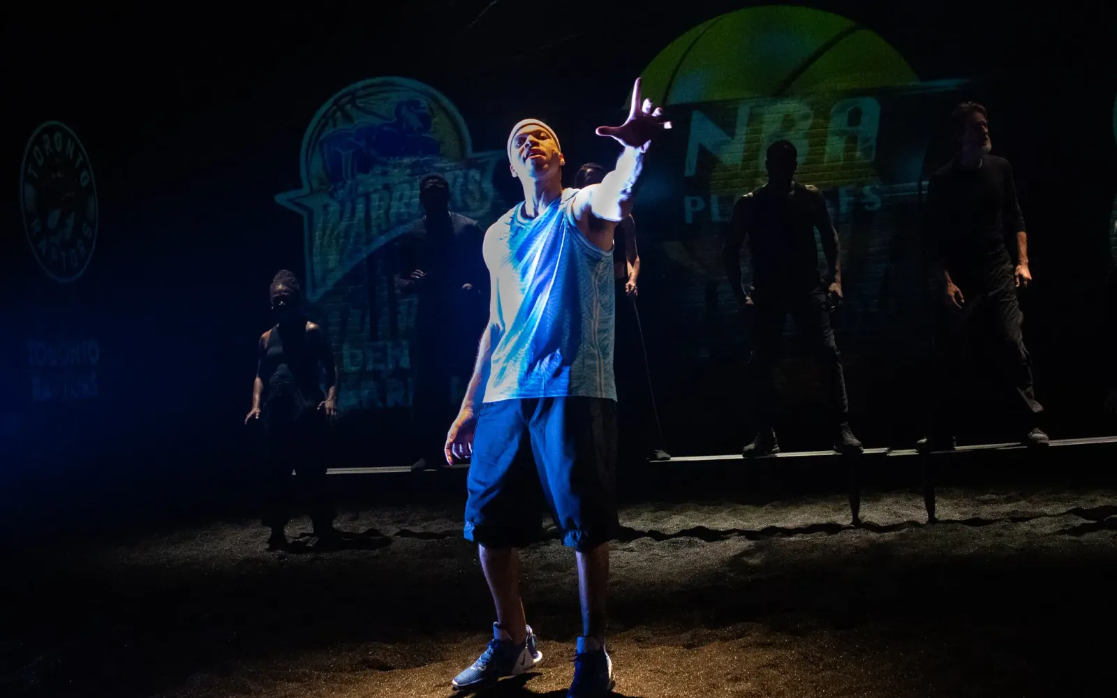 A man in a blue basketball jersey and shorts stands center stage in a spotlight, with a few figures in shadow in the background.
