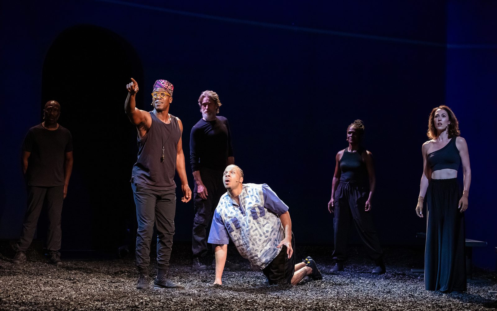 Jason Bowen in glasses and a crown kufi hat points in the distance as Mister Fitzgerald rises from his knees, and the other actors watch.