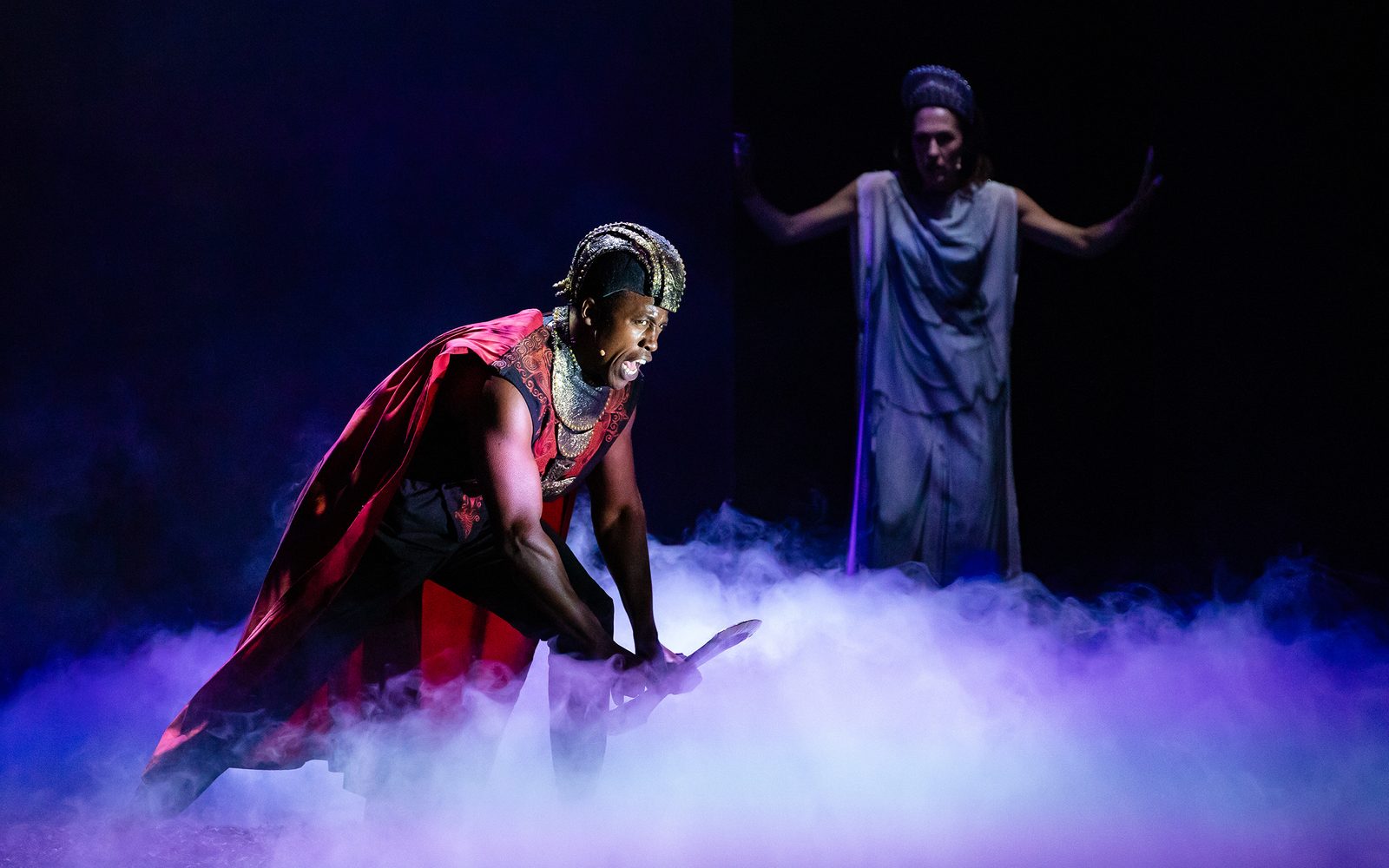 Jason Bowen in bronze armor picks up an axe while Kelley Curran in a toga watches behind him.