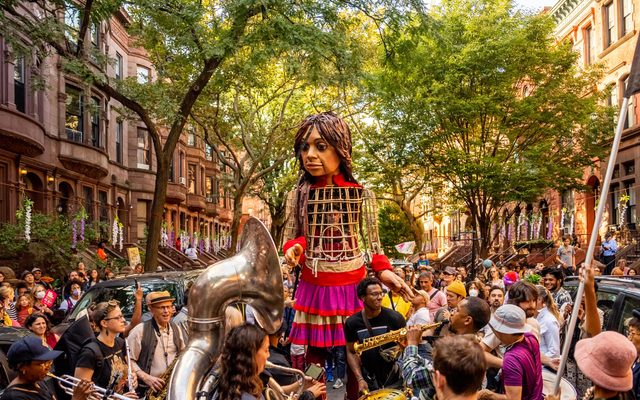 Little Amal walks down a tree-lined street behind a group of brass players.