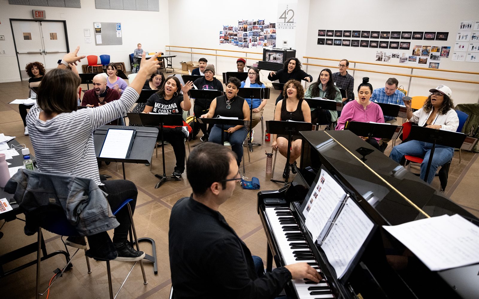 The Music Supervisor lifts her hands as the company sings from their music stands.