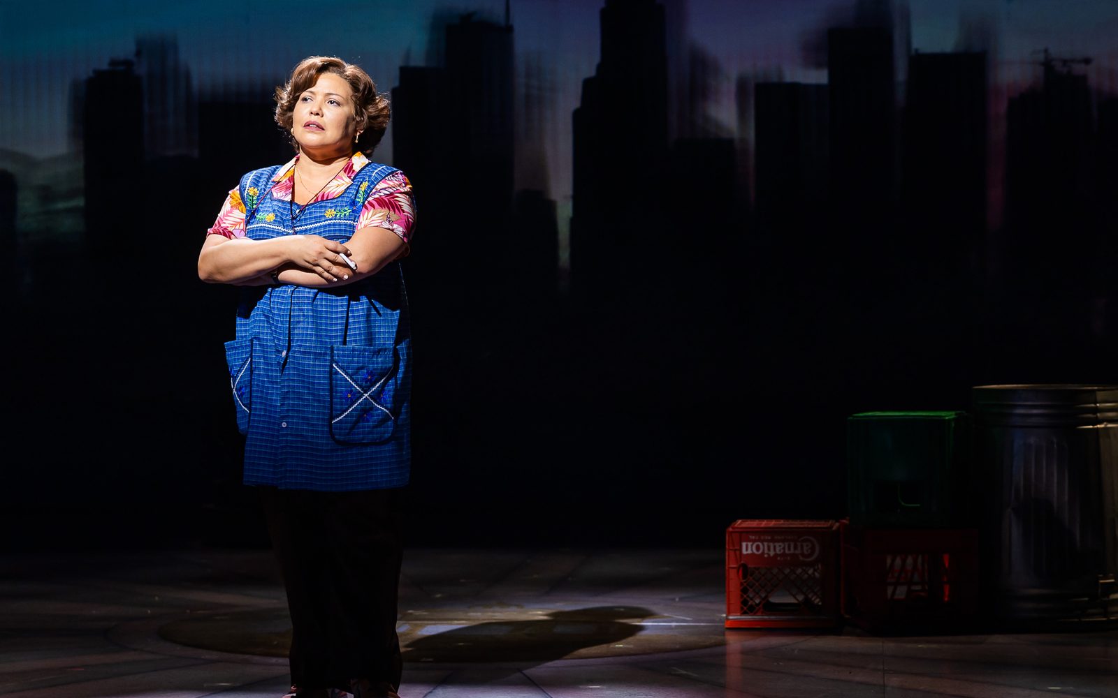 Justina Machado in her sewers uniform stands with her arms crossed and holding a cigarette against a gray backdrop.
